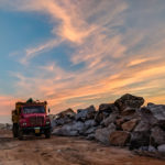 A truck hauling junk on a rocky beach at sunset.
